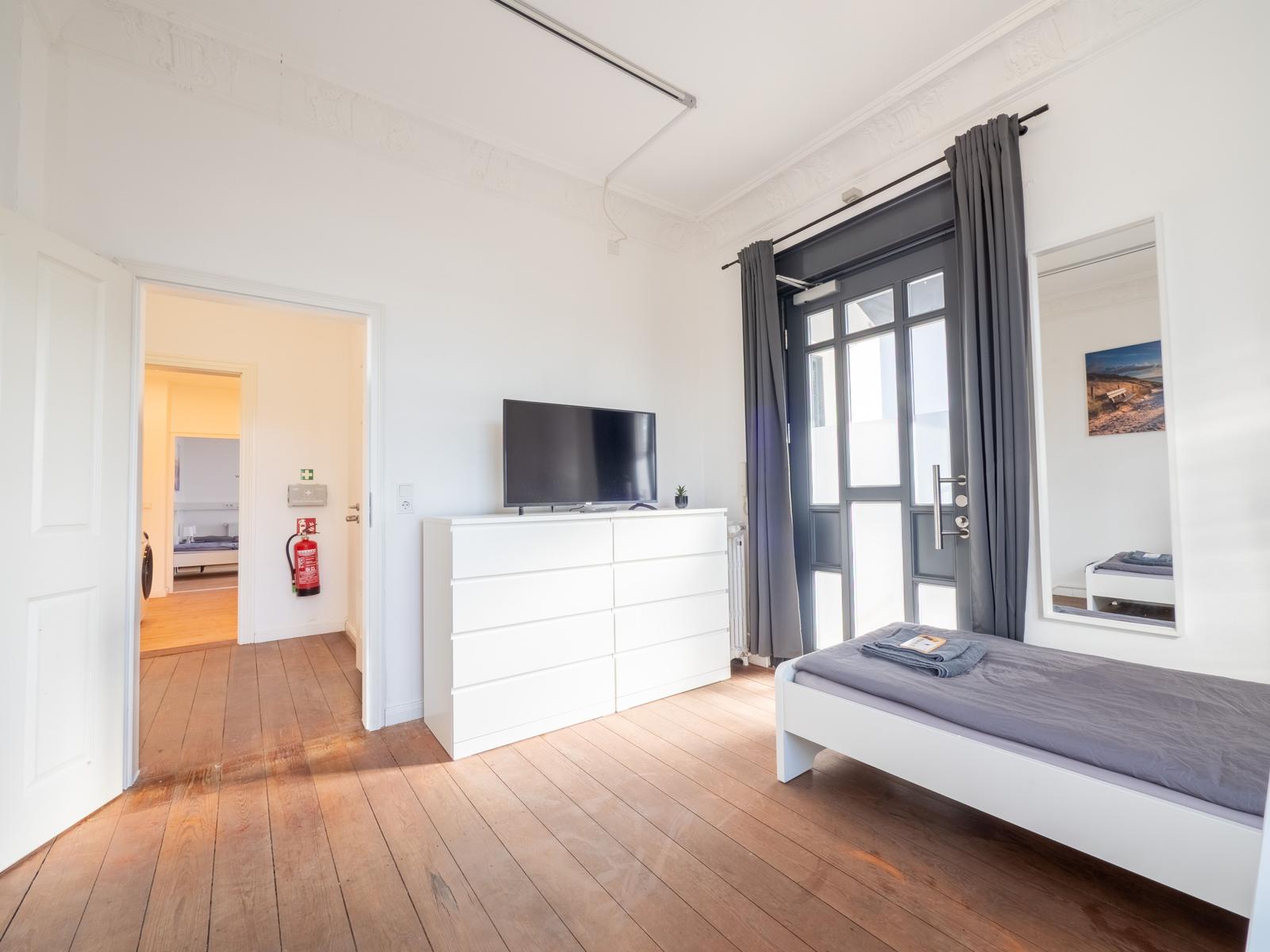 Schlafzimmer mit Bett, TV und Fenster. Holzboden und weiße Wände.