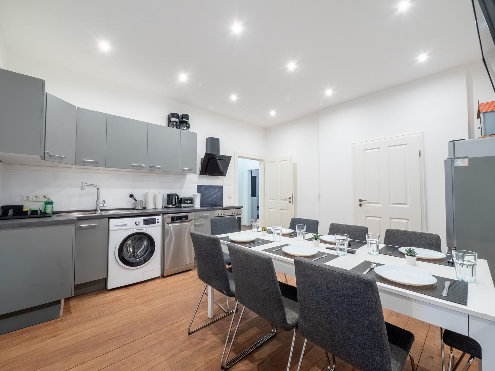 Küche mit Essbereich, Waschmaschine und Kühlschrank. Holzboden und graue Möbel.