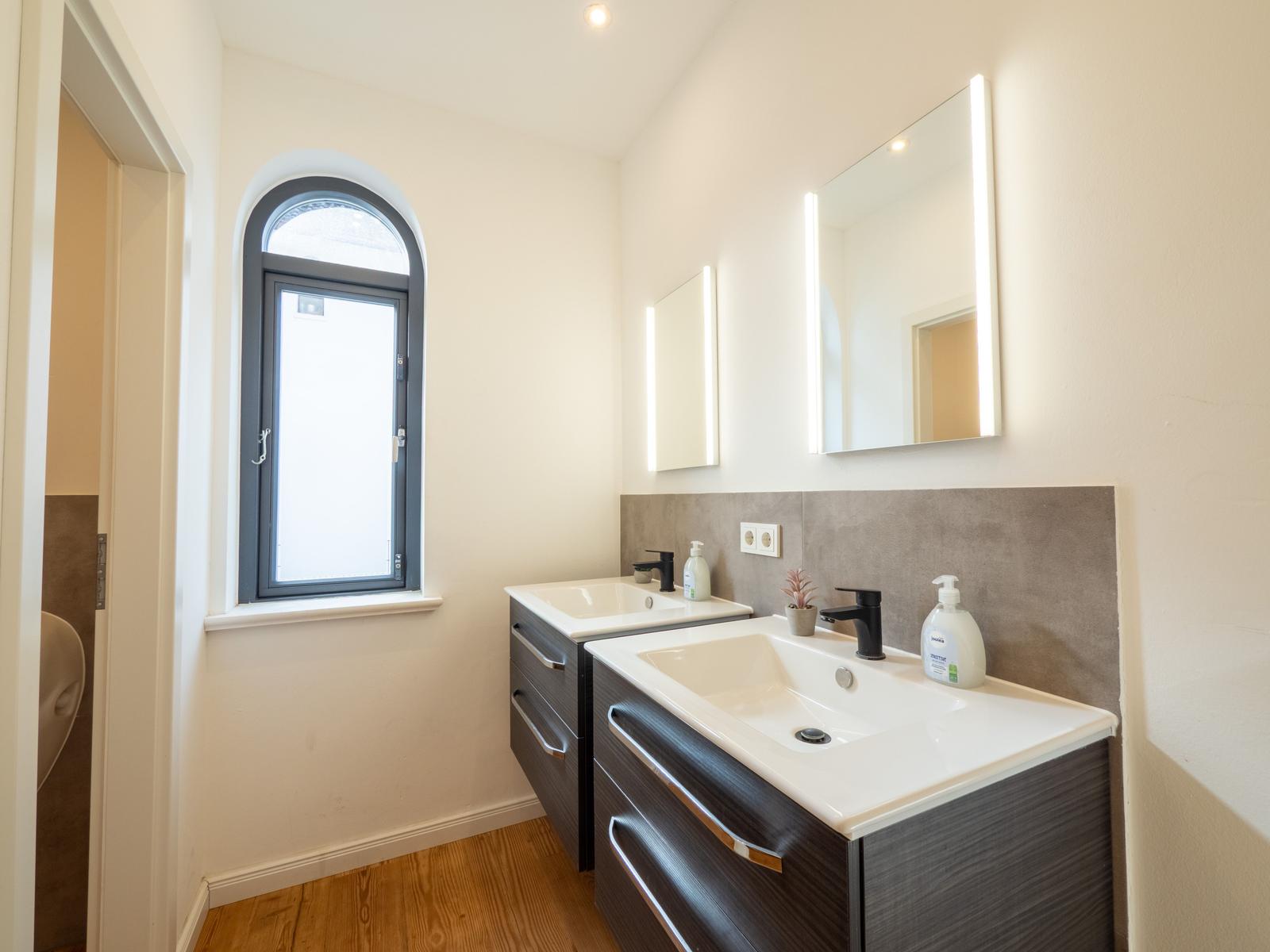 Badezimmer mit zwei Waschbecken, beleuchtetem Spiegel und Badezimmerfenster.