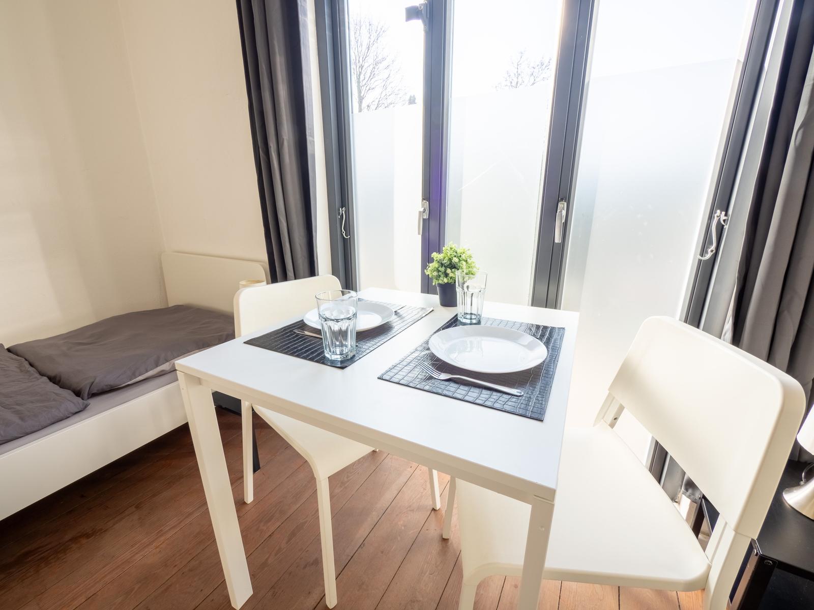 Essbereich mit Tisch, zwei Stühlen und Fensterblick. Hintergrund: Bett mit grauer Bettwäsche.
