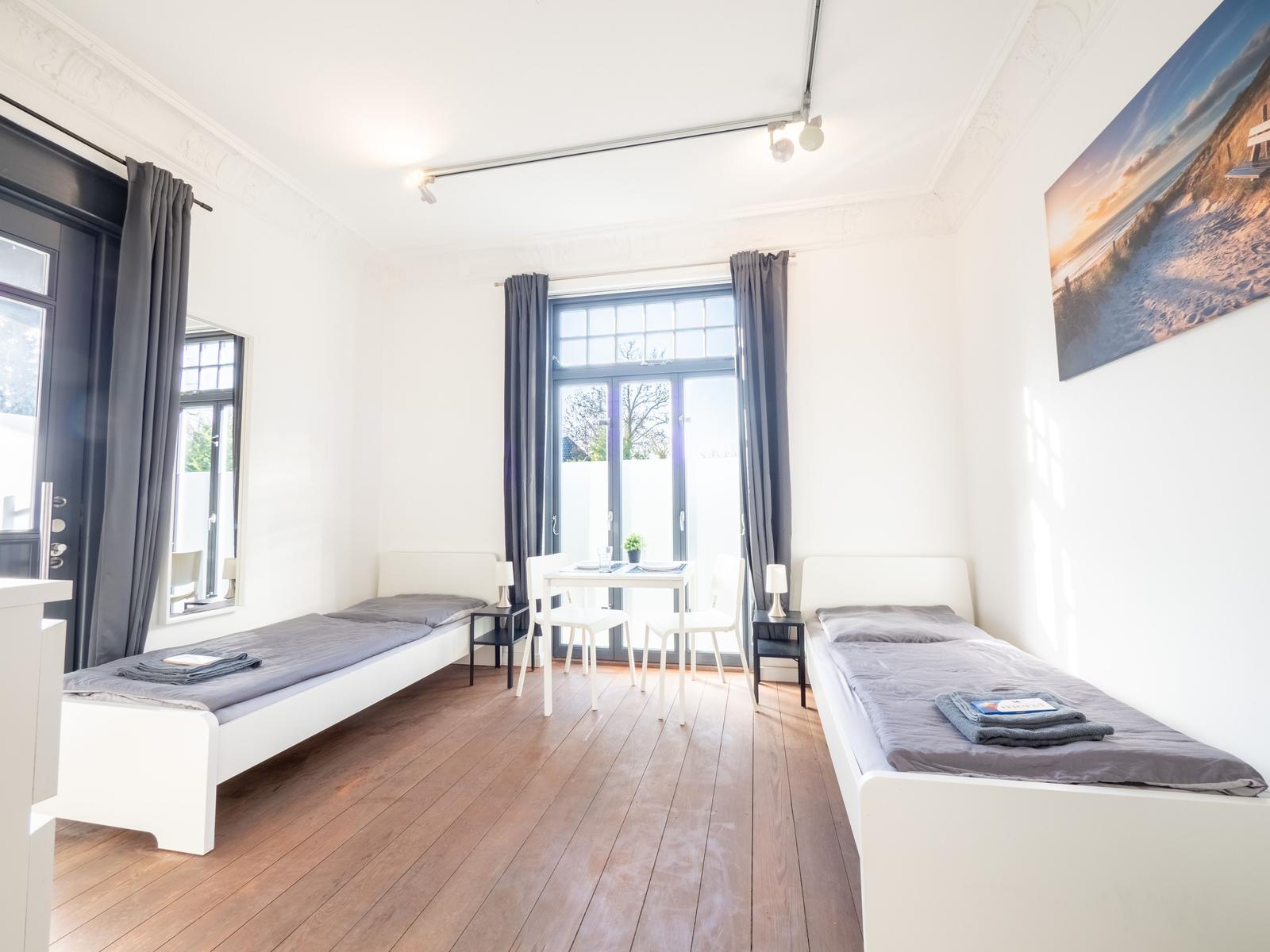 Zwei Betten, Tisch mit Stühlen, Fenster und Wandbild im Raum.