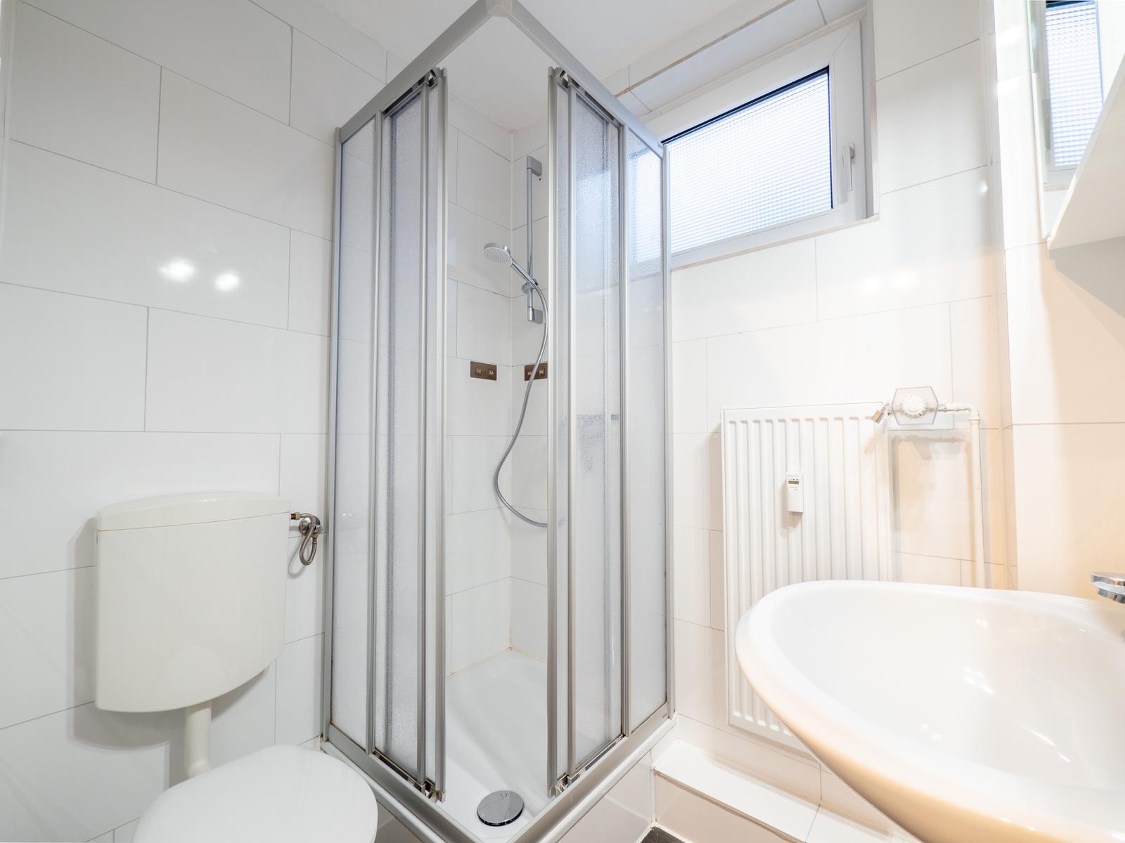 Badezimmer mit Dusche, Waschbecken und Toilette. Weiße Fliesen und Fenster.