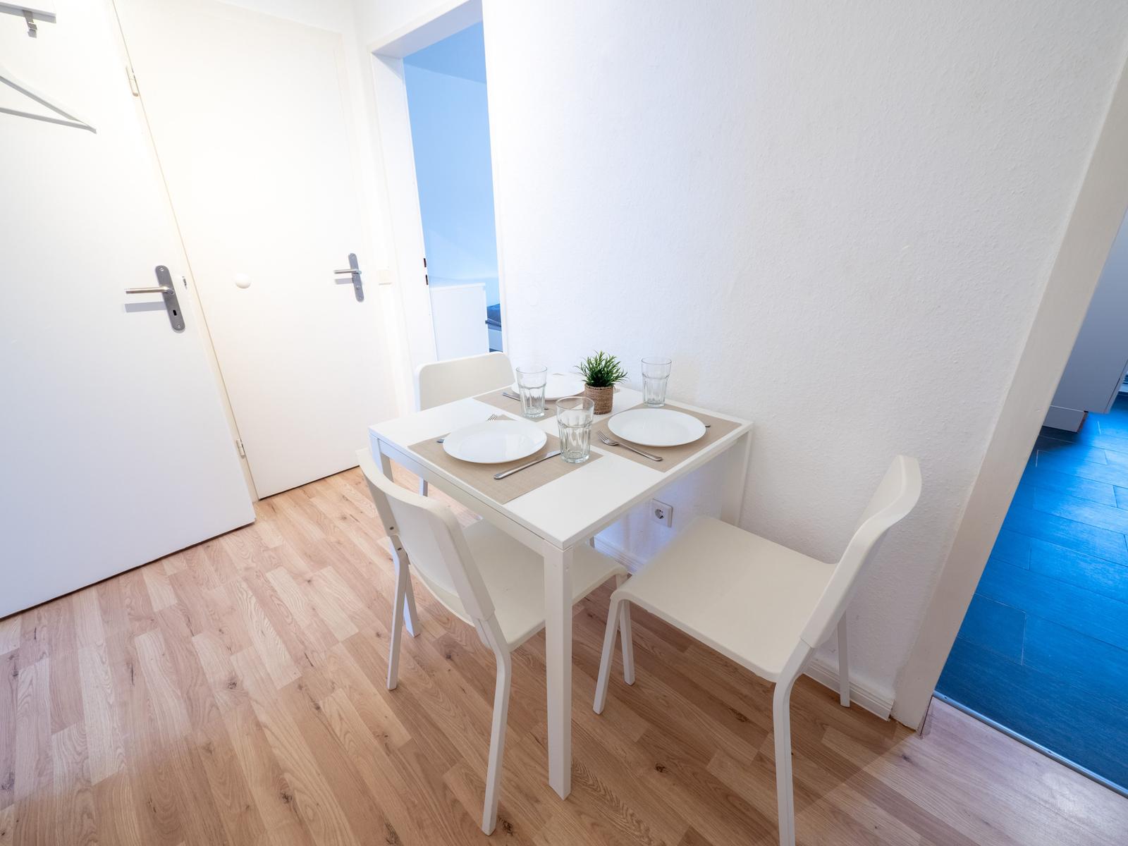 Esstisch mit vier Stühlen, Tischdecke und Geschirr. Holzboden und weiße Wände.