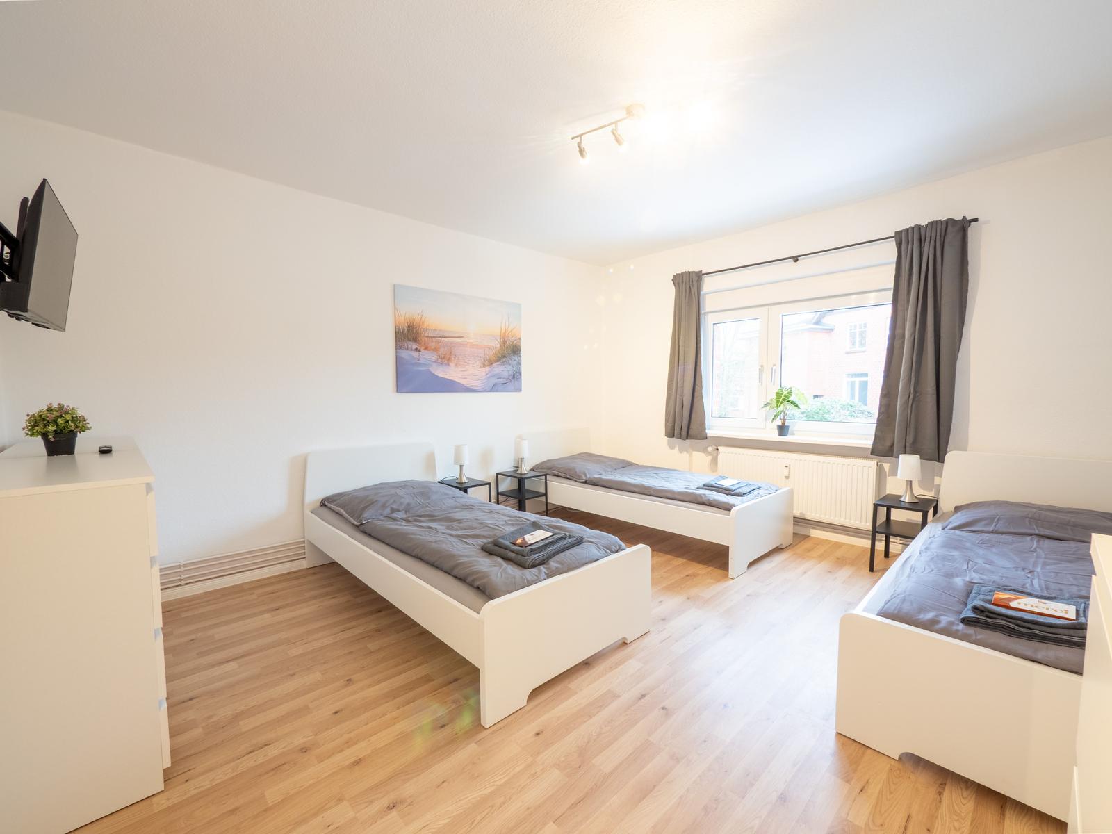 Drei Einzelbetten, Fenster mit Vorhängen, Wandbild, Fernseher und Holzboden.