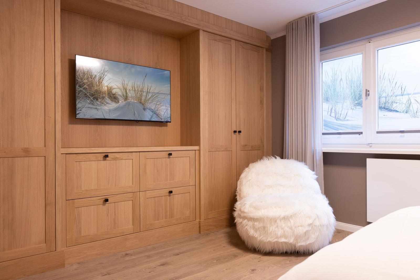 Schlafzimmer mit Holzmöbeln, Fernseher und flauschigem Sitzkissen.