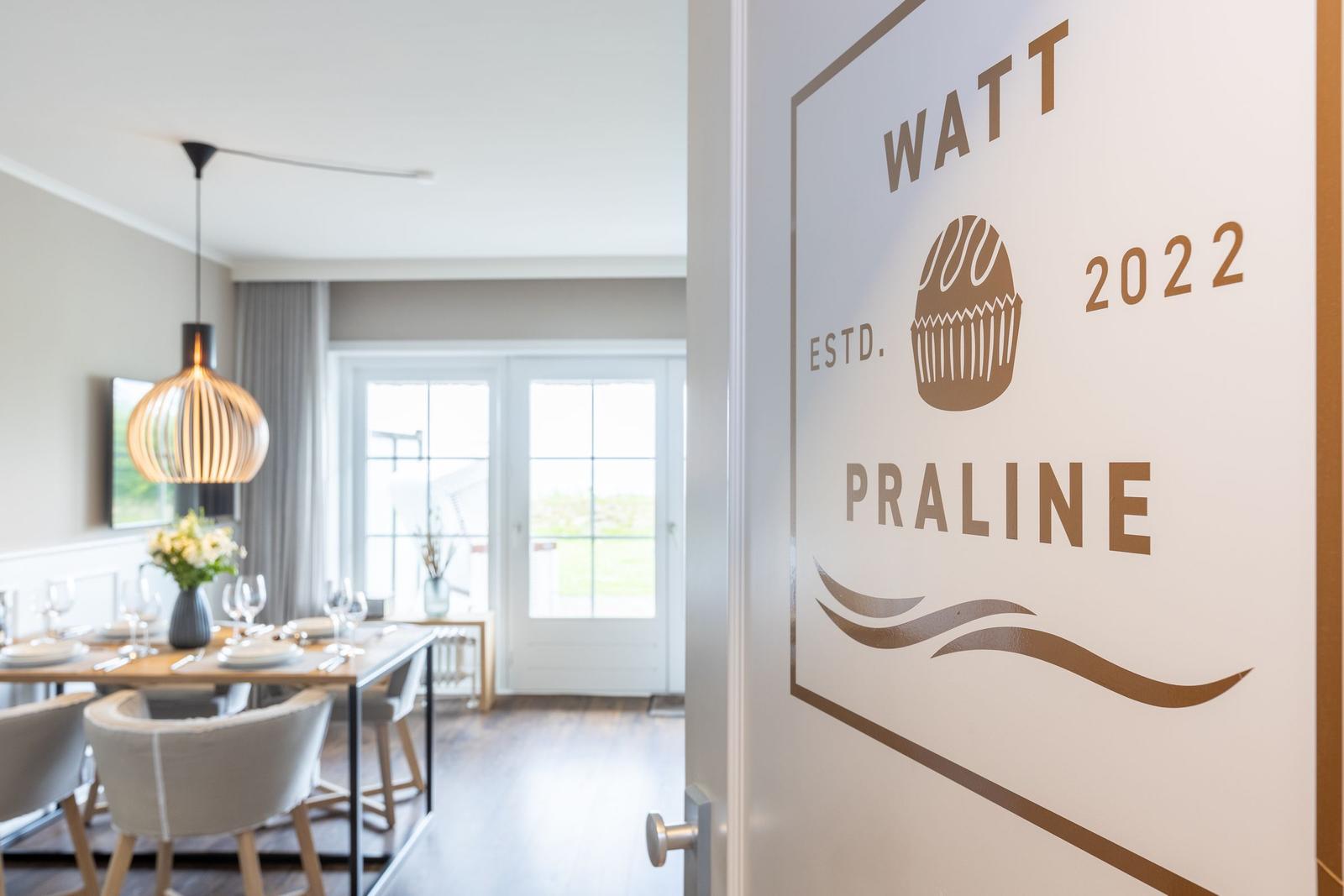 Tür mit Logo WATT PRALINE ESTD. 2022. Im Hintergrund Essbereich mit Tisch und Stühlen.