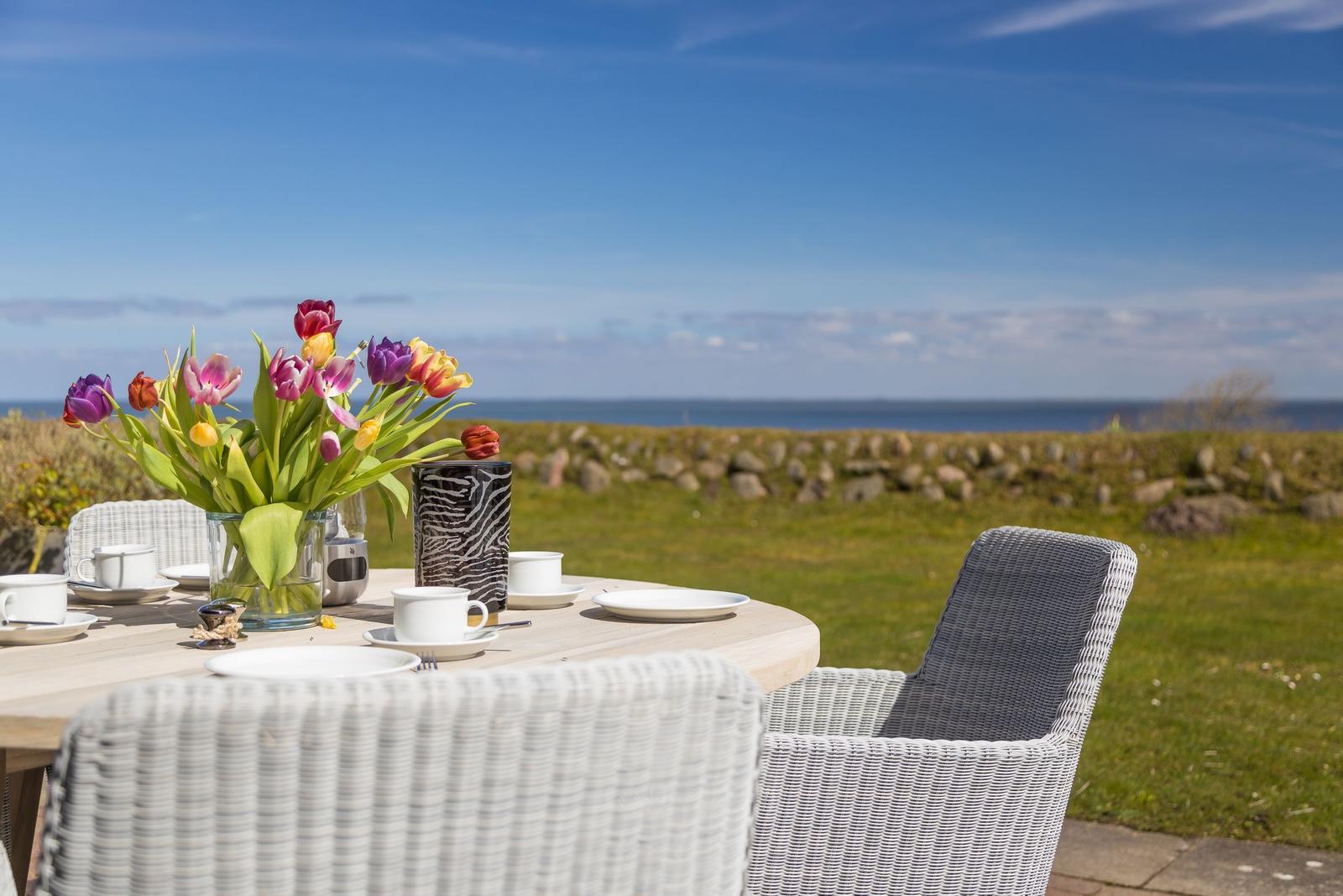 Rundtisch mit Blumen und Tassen im Freien, Blick auf Meer und Gras.
