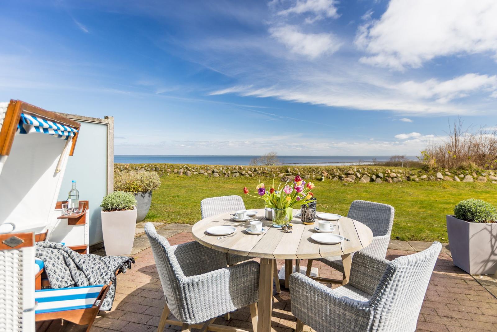 Terrasse mit Tisch, Stühlen und Meerblick. Strandkorb und Blumenarrangement sichtbar.