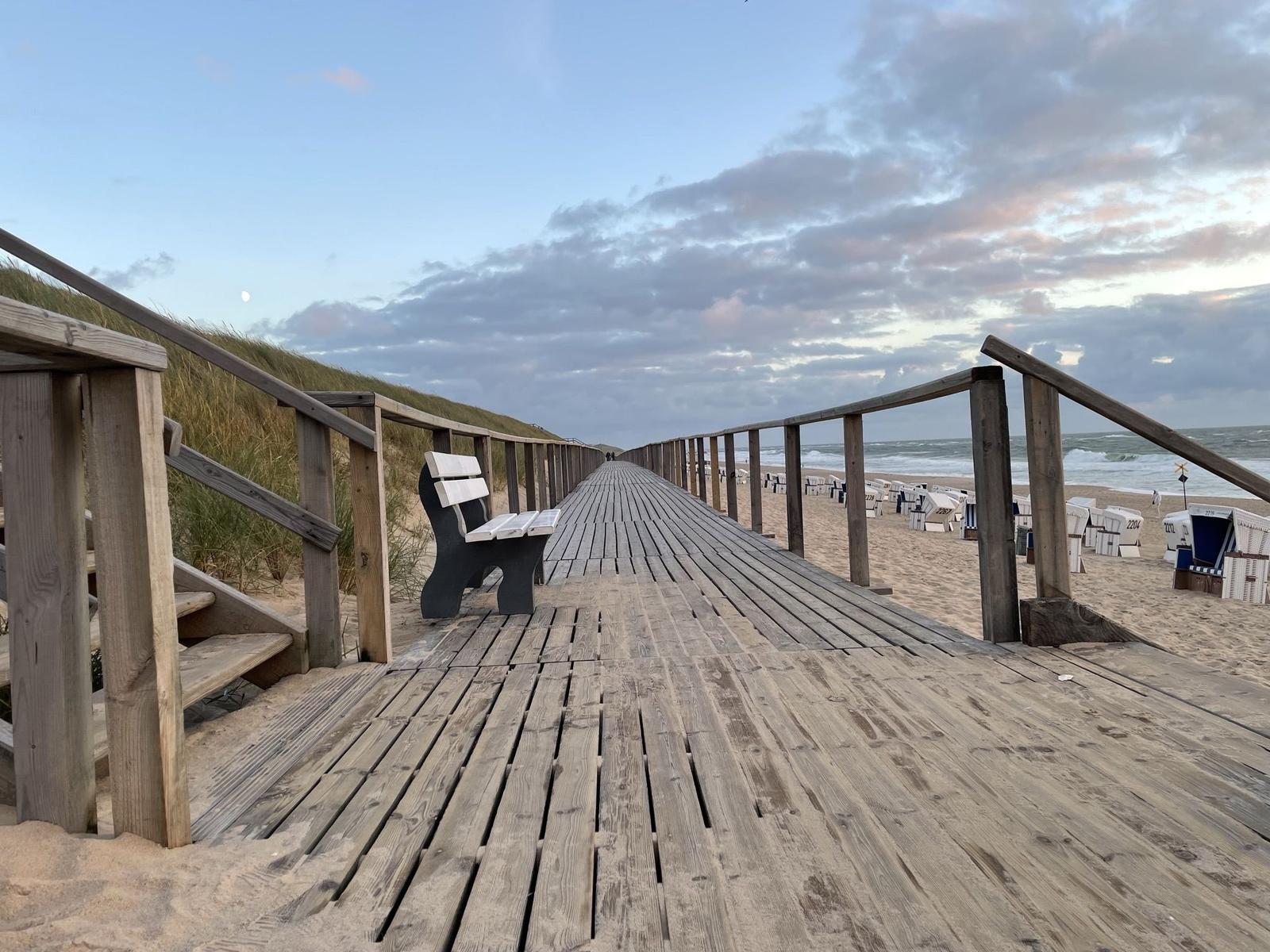 Holzpromenade führt zum Strand mit Strandkörben und Wellen.
