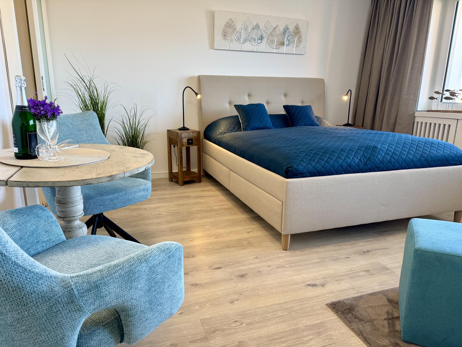 Schlafzimmer mit Bett, Tisch und Stühlen. Blaue Kissen und Wandbild.