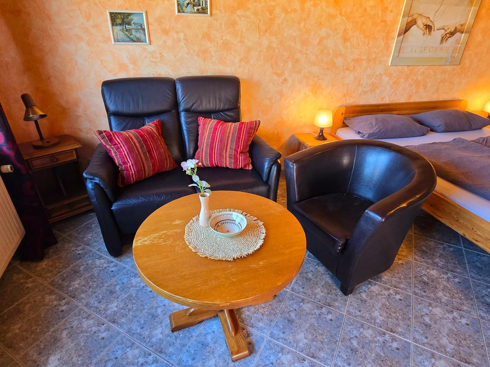 Zimmer mit Sofa, Sessel, Tisch und Bett. Wände orange, Boden Fliesen.