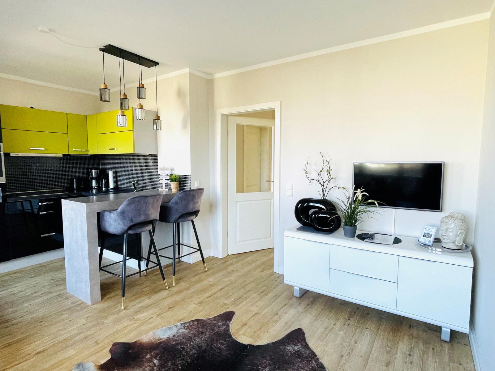 Küche mit gelben Schränken und Essbereich. Wohnbereich mit Fernseher und weißen Möbeln.