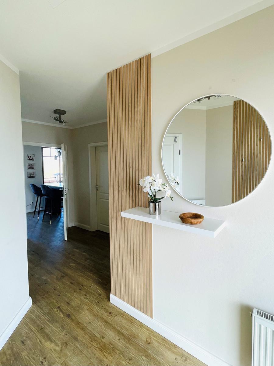 Hausflur mit runden Spiegel, Holzpaneelen und Schrank. Blick in die Küche mit Stühlen.