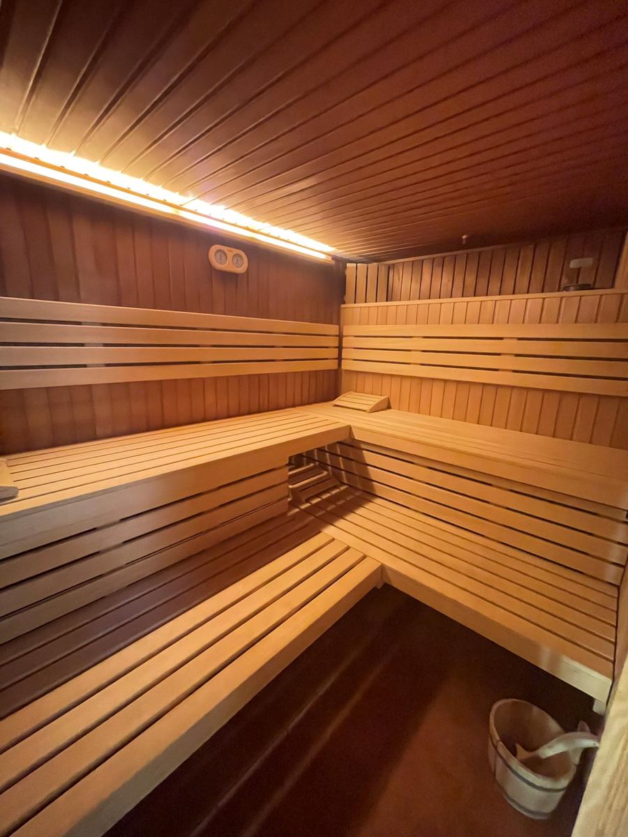 Sauna mit Holzbanken und Beleuchtung. Eimer und Thermostat sichtbar.