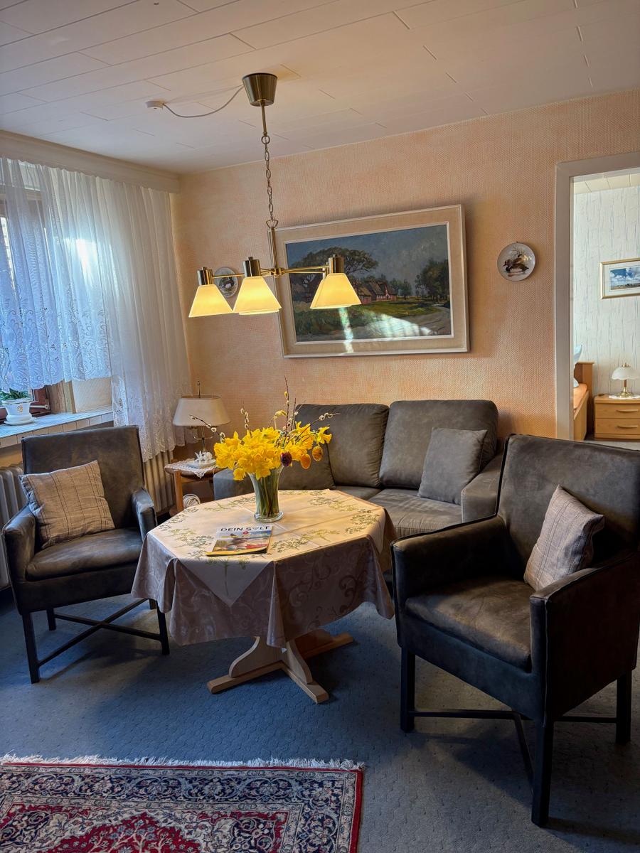 Wohnzimmer mit Sofa, zwei Sesseln, Tisch und Blumen. Wandbild und Deckenleuchte sichtbar.