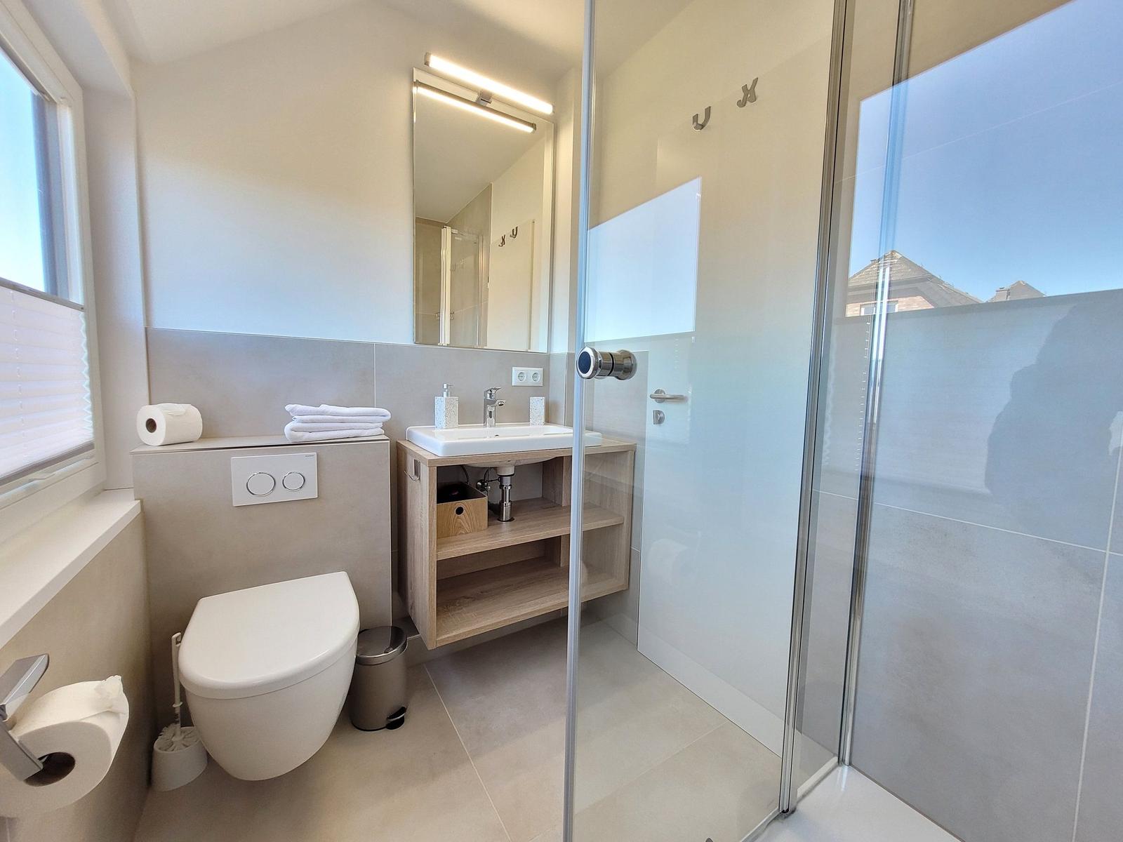 Badezimmer mit Dusche, Waschbecken und Toilette. Fenster mit Jalousie.