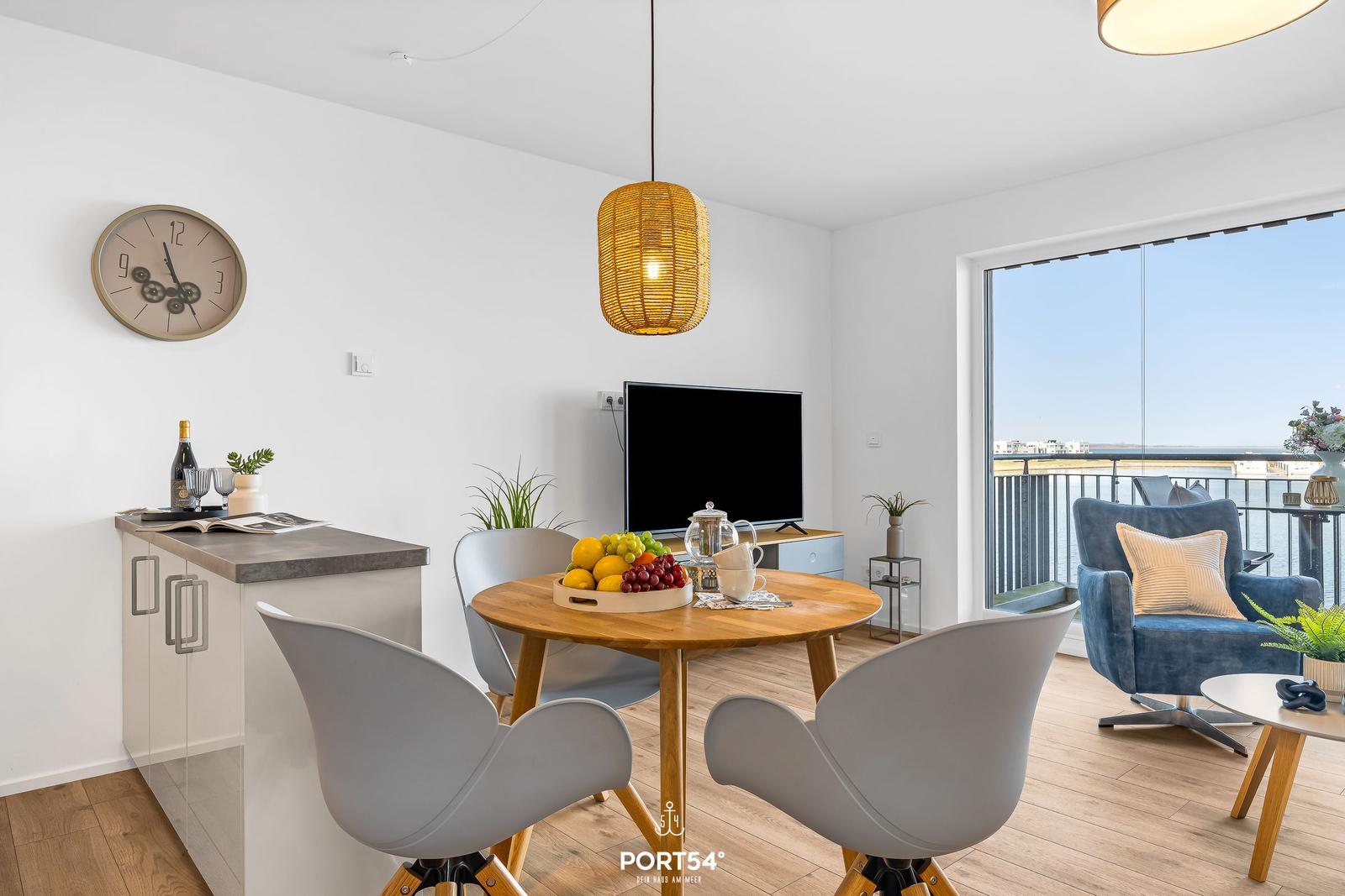 Essbereich mit Tisch, Stühlen und Fernseher. Balkon mit Sitzgelegenheit und Blick auf Wasser.