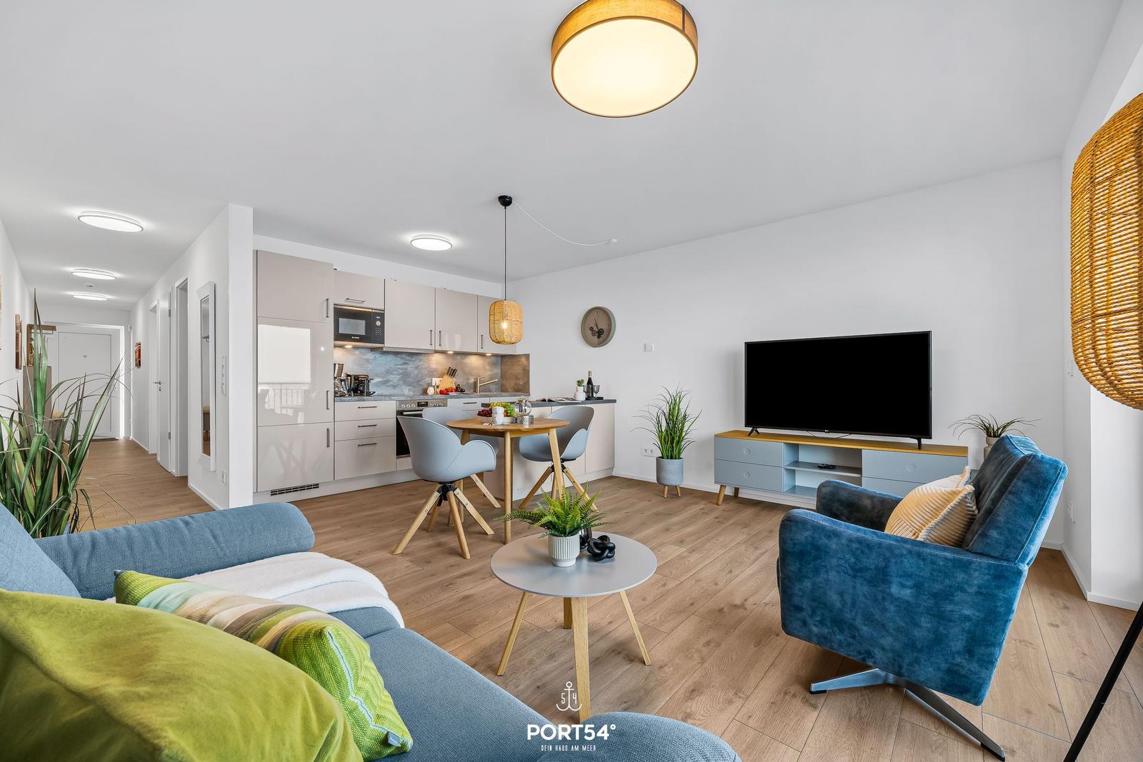 Wohnbereich mit Küche, Essplatz und TV. Holzboden und blaue Sitzgelegenheiten.