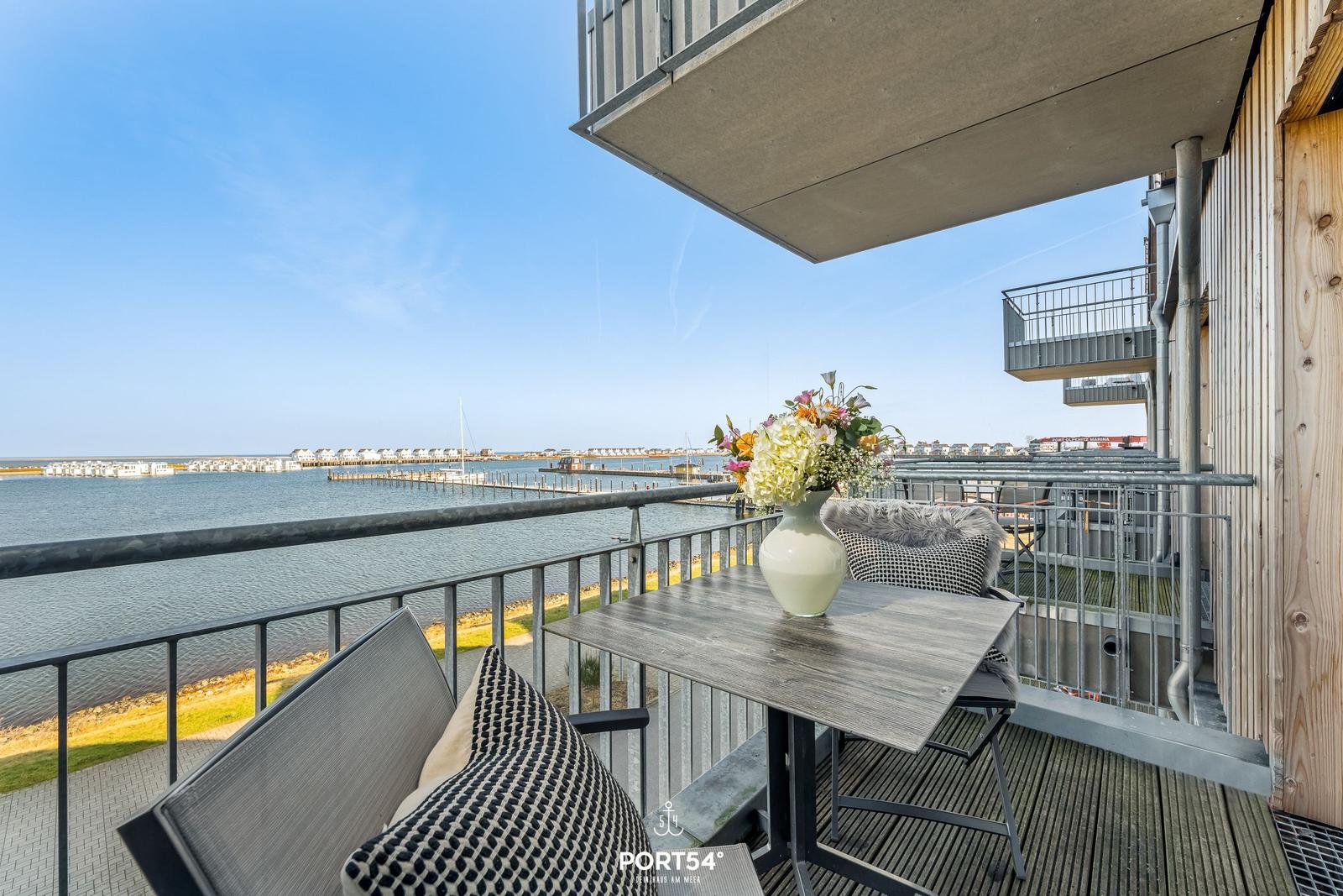 Terrasse mit Tisch, Stühlen und Blumenvase. Blick auf Wasser und Steg.