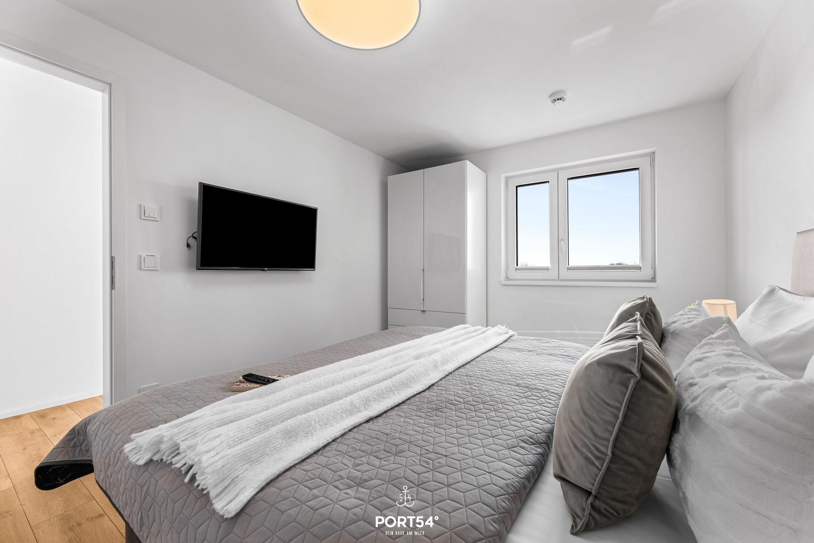 Schlafzimmer mit Bett, TV und Fenster. Holzboden und weißer Kleiderschrank.