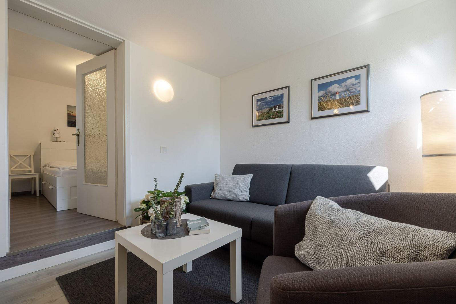 Wohnzimmer mit Sofa, Tisch und Bildern. Tür führt in Schlafzimmer.