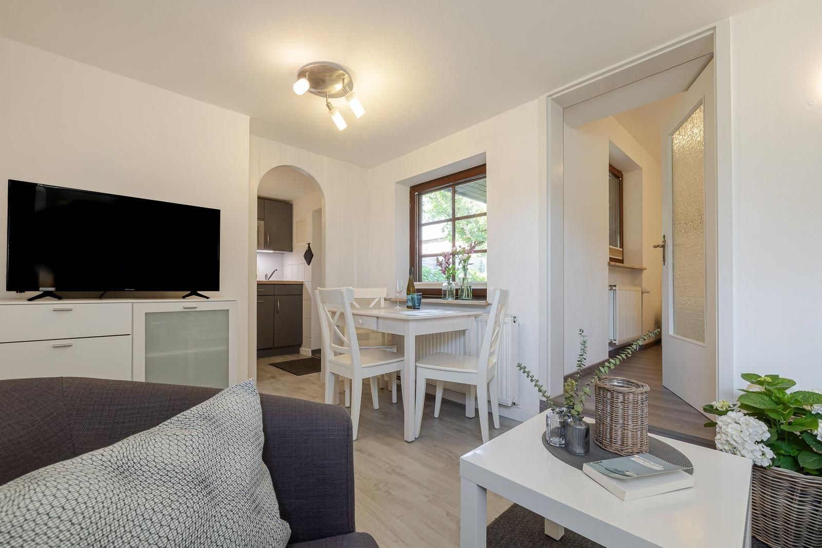 Wohn- und Essbereich mit TV, Tisch und Stühlen. Küche und Tür sichtbar.