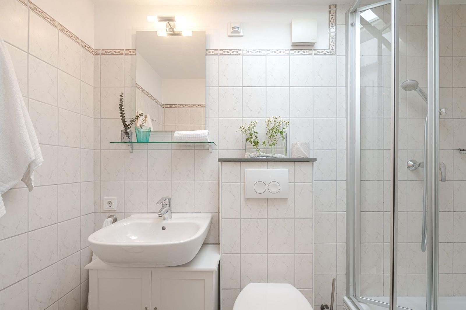 Badezimmer mit Dusche, Waschbecken und Toilette. Weiße Fliesen und Glasregal.