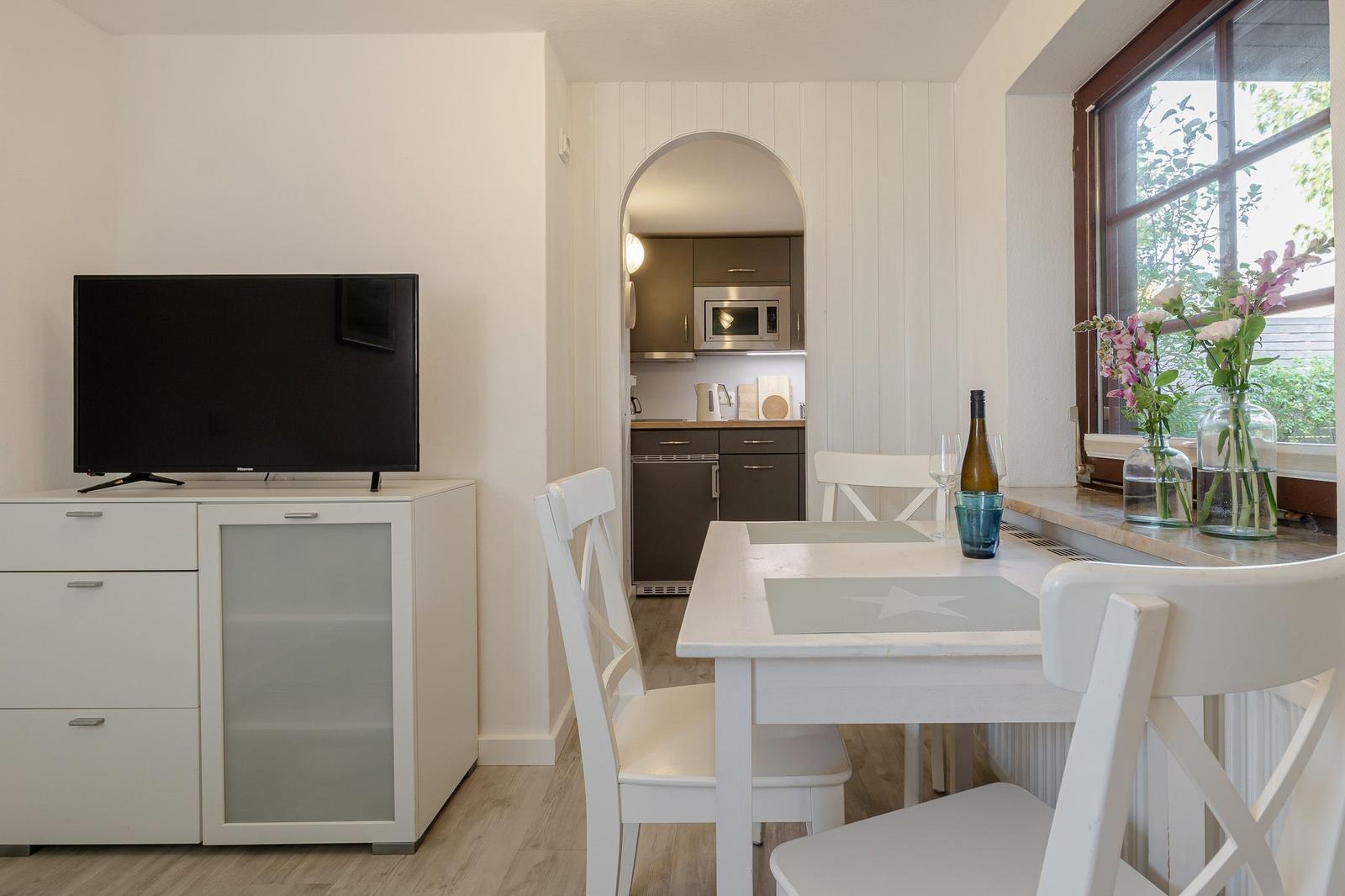 Essbereich mit Tisch, Stühlen und Fernseher. Küche im Hintergrund sichtbar.