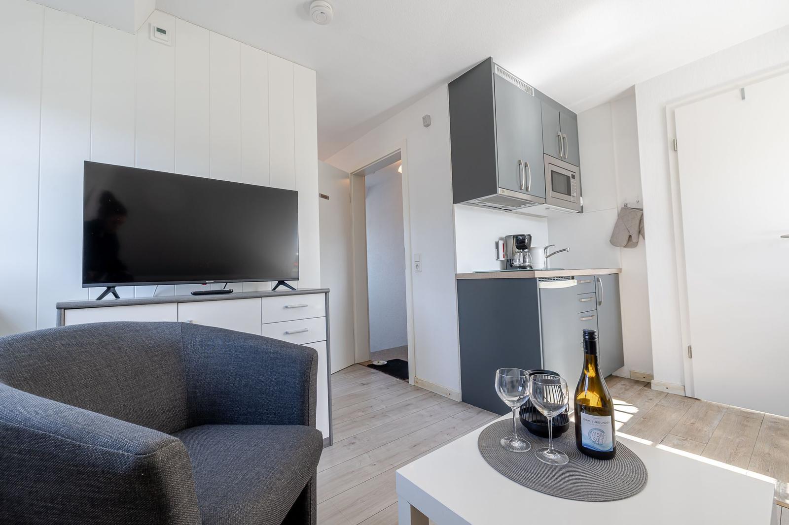 Wohnbereich mit TV, Küchenzeile und Sitzgelegenheit. Flur führt in das Schlafzimmer.