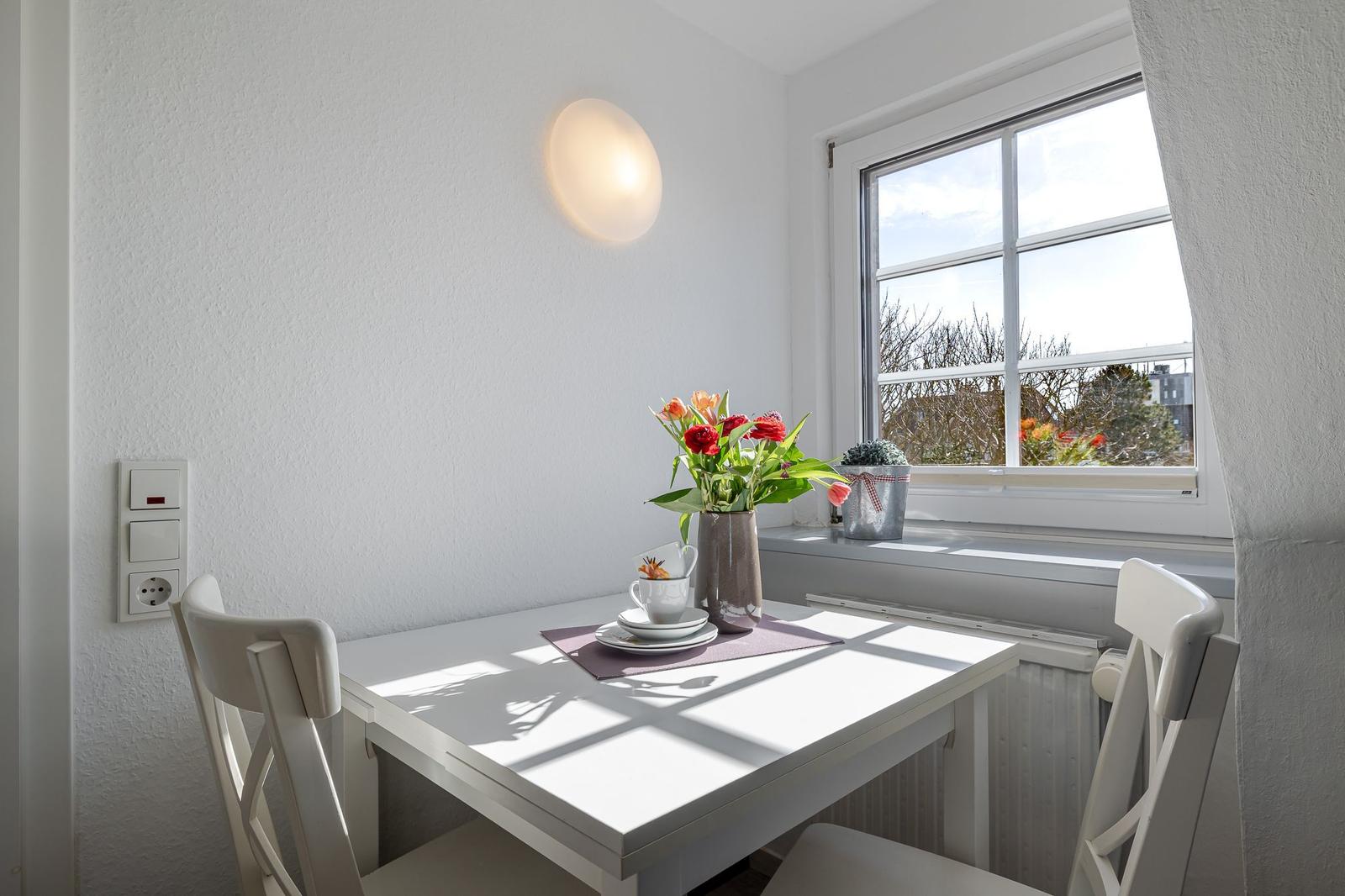 Ein weißer Tisch mit zwei Stühlen, Blumen und einer Tasse vor einem Fenster.