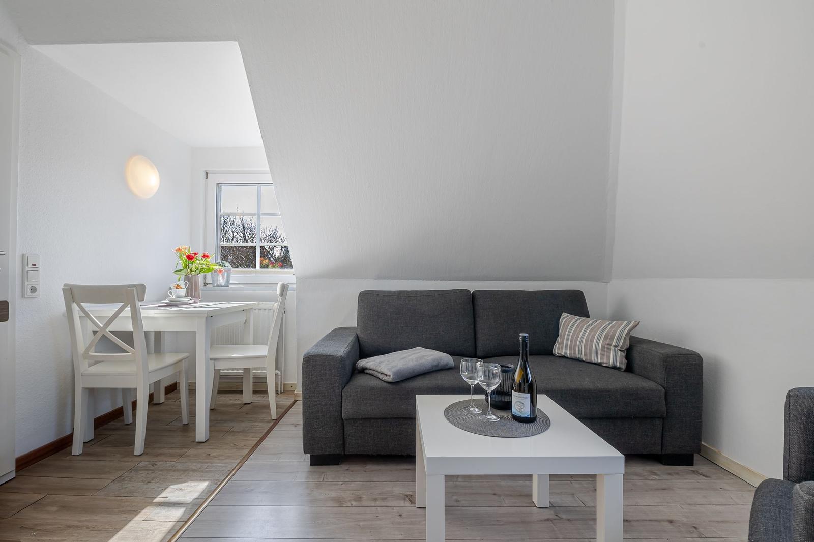 Wohnzimmer mit grauem Sofa, weißem Tisch und Stühlen, Flur mit Tisch und Blumen.