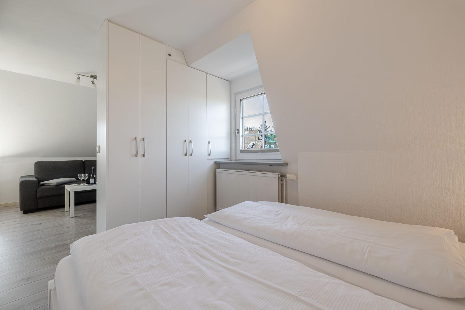 Schlafzimmer mit Bett, Kleiderschrank und Fenster. Im Hintergrund Wohnbereich mit Sofa.