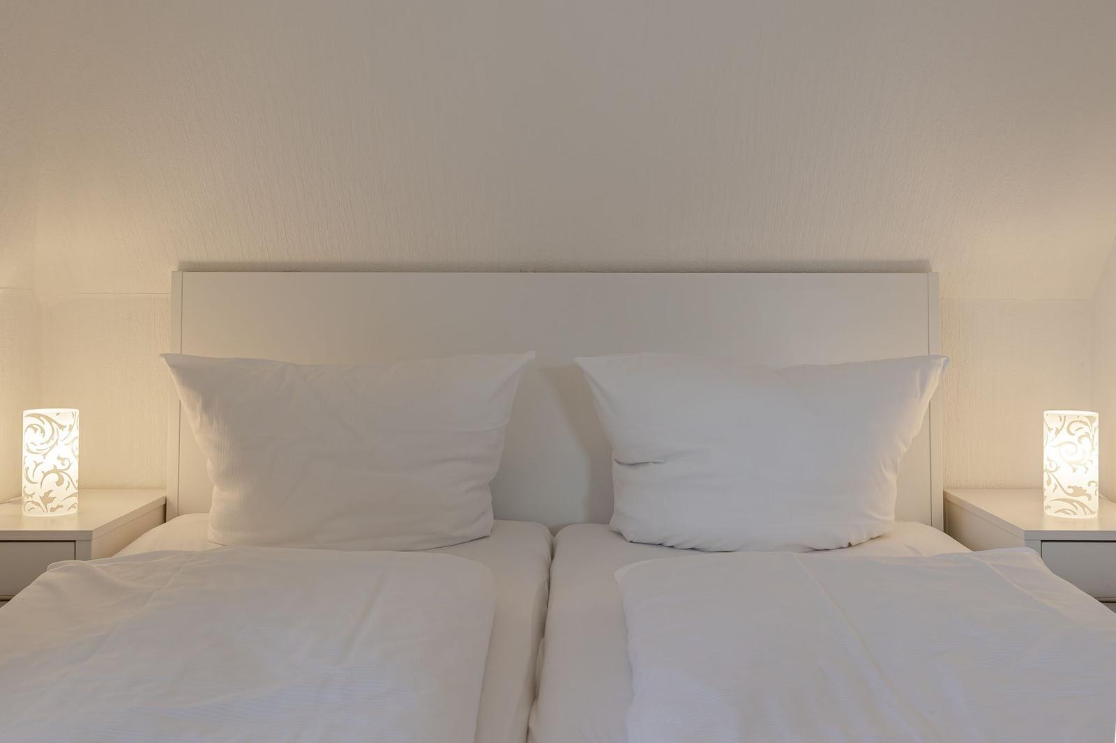 Zwei Betten mit weißen Kissen und Bettwäsche. Beide Nachttische haben eine Lampe.