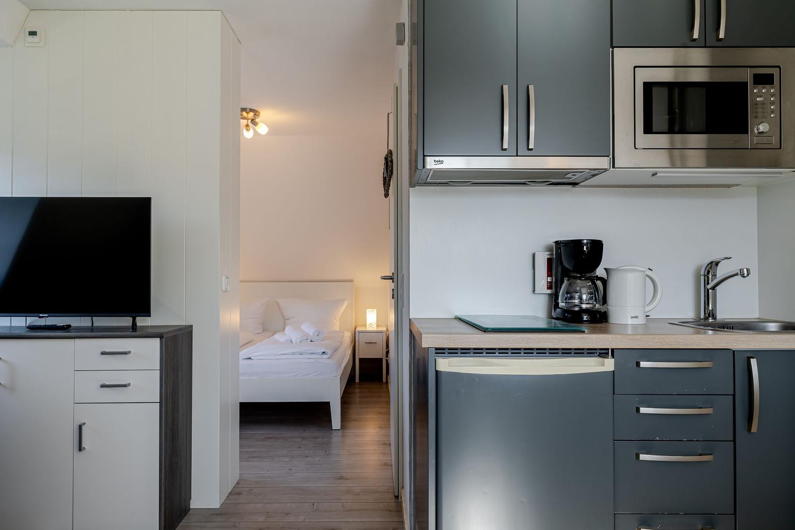 Küche mit Kühlschrank, Mikrowelle und Kaffeemaschine. Blick in Schlafzimmer mit Bett und Fernseher.