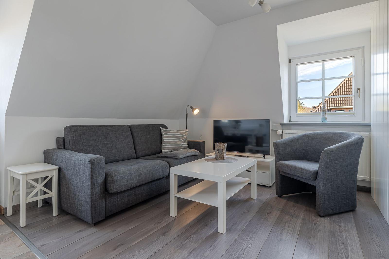 Wohnzimmer mit Sofa, Sessel, Couchtisch und TV. Holzboden und Fenster mit Blick.