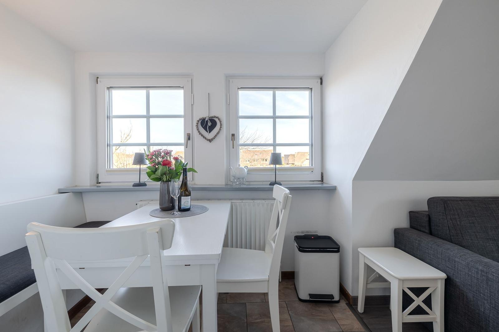 Essbereich mit weißem Tisch, Stühlen und Fenstern. Sofa und Fensterbank sichtbar.