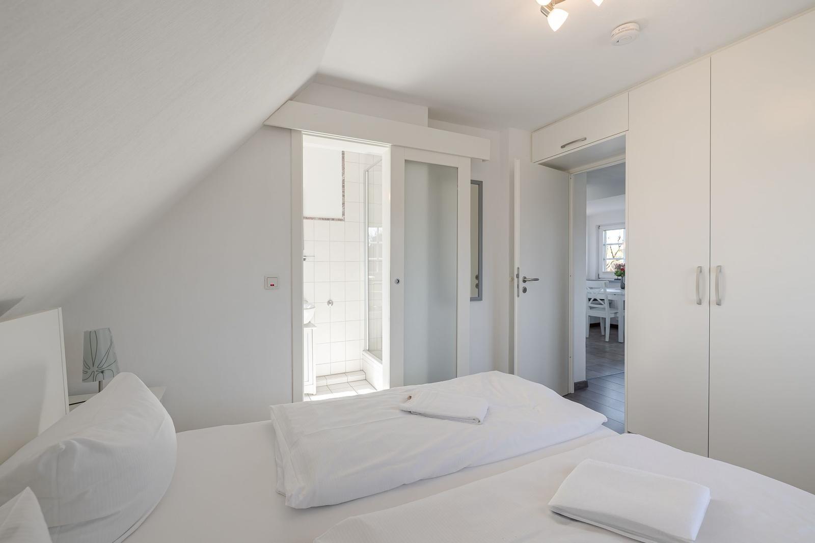 Schlafzimmer mit Dachschräge, weißem Bett und Anschluss zu Bad und Wohnbereich.