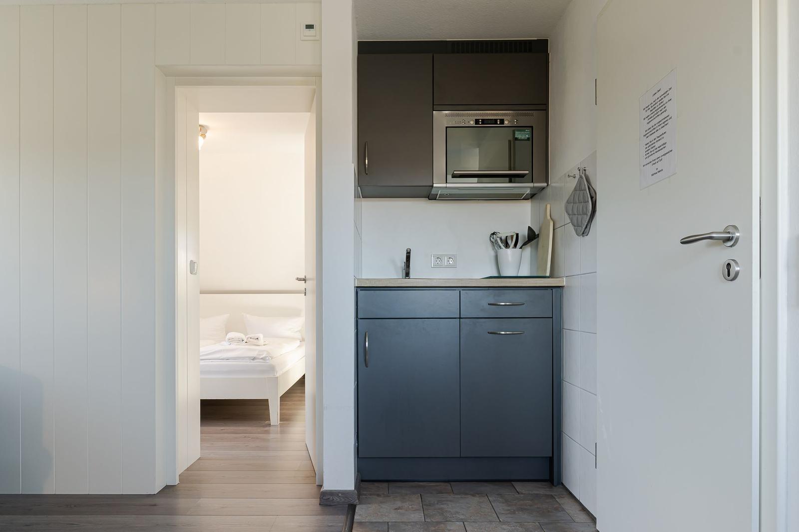 Küche mit Dunstabzug und Mikrowelle. Schlafzimmer mit weißem Bett im Hintergrund.