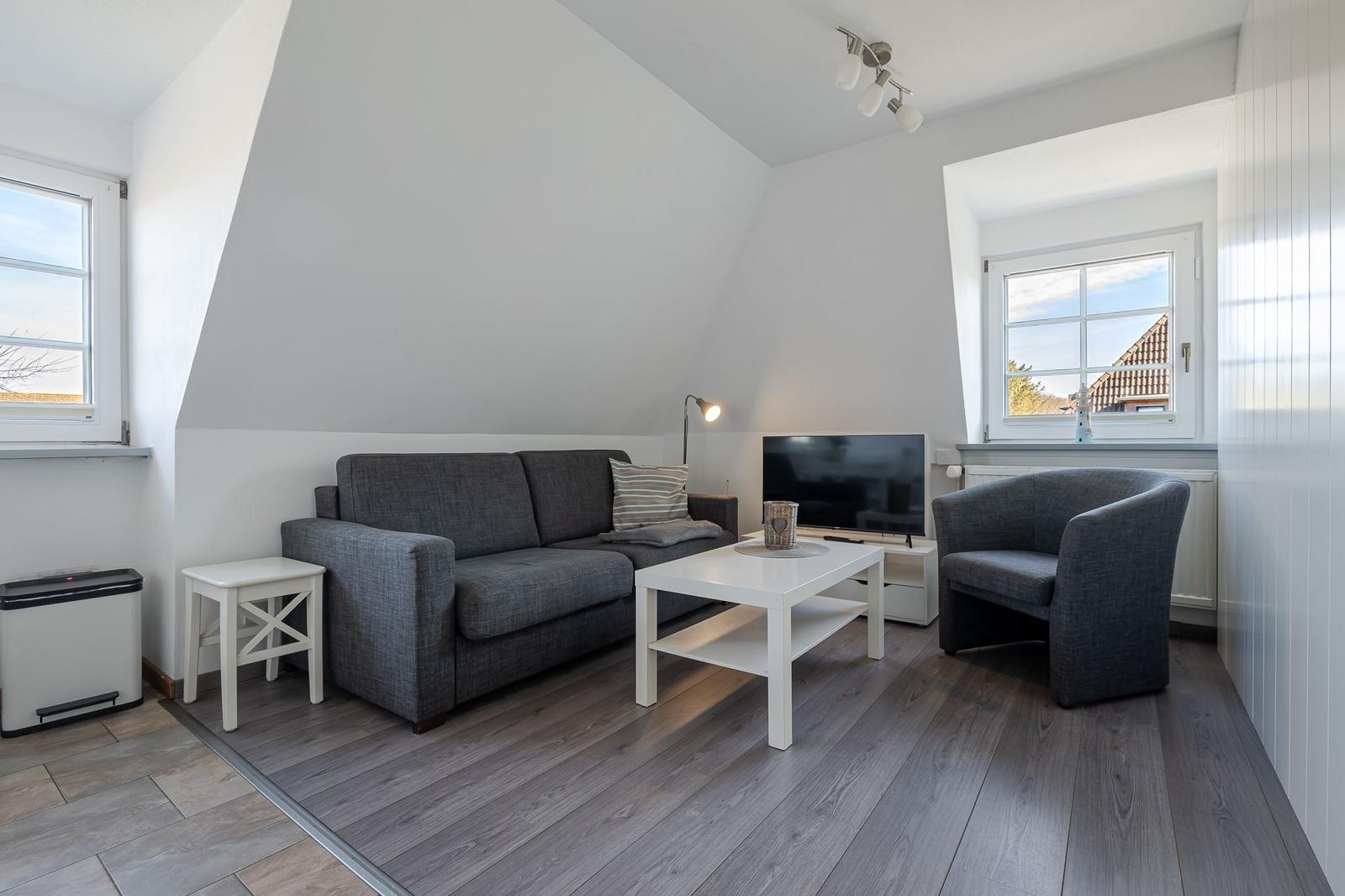 Wohnzimmer mit grauem Sofa, weißen Möbeln und Holzboden. Fenster bieten Blick auf Dachziegel.