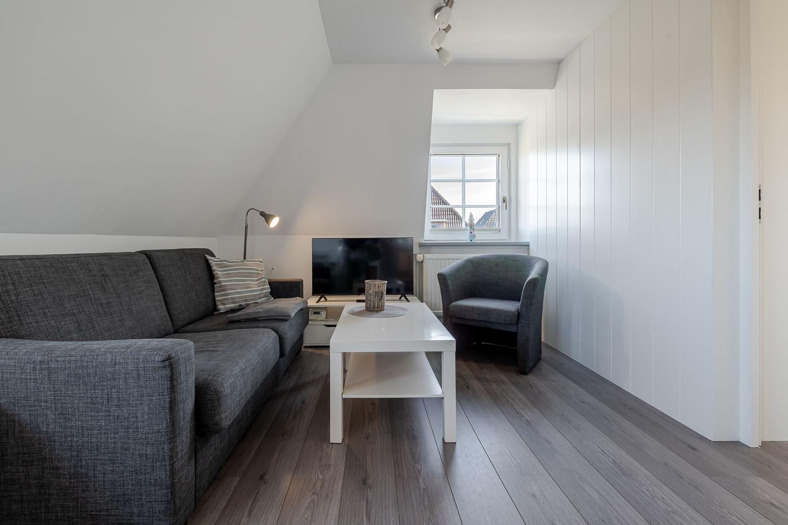 Wohnzimmer mit Sofa, Sessel, Couchtisch und Fernseher. Holzboden und weiß gestrichene Wände.