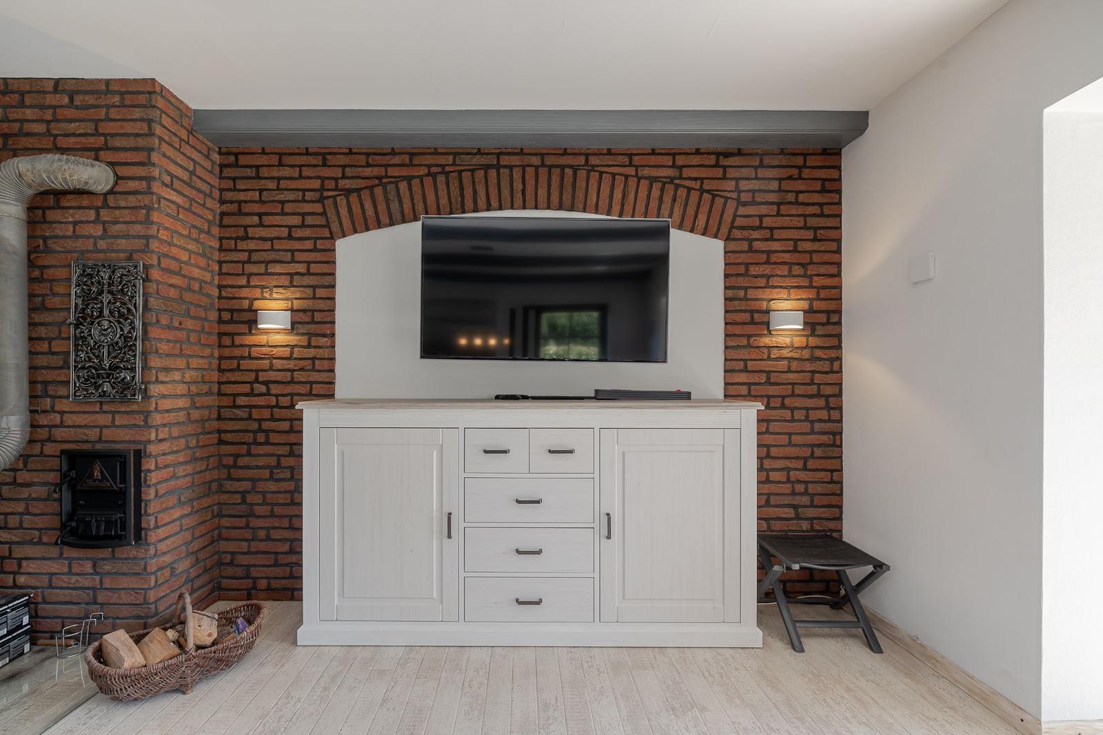Wohnzimmer mit Kamin, Fernseher und weißen Schränken an Backsteinwand.