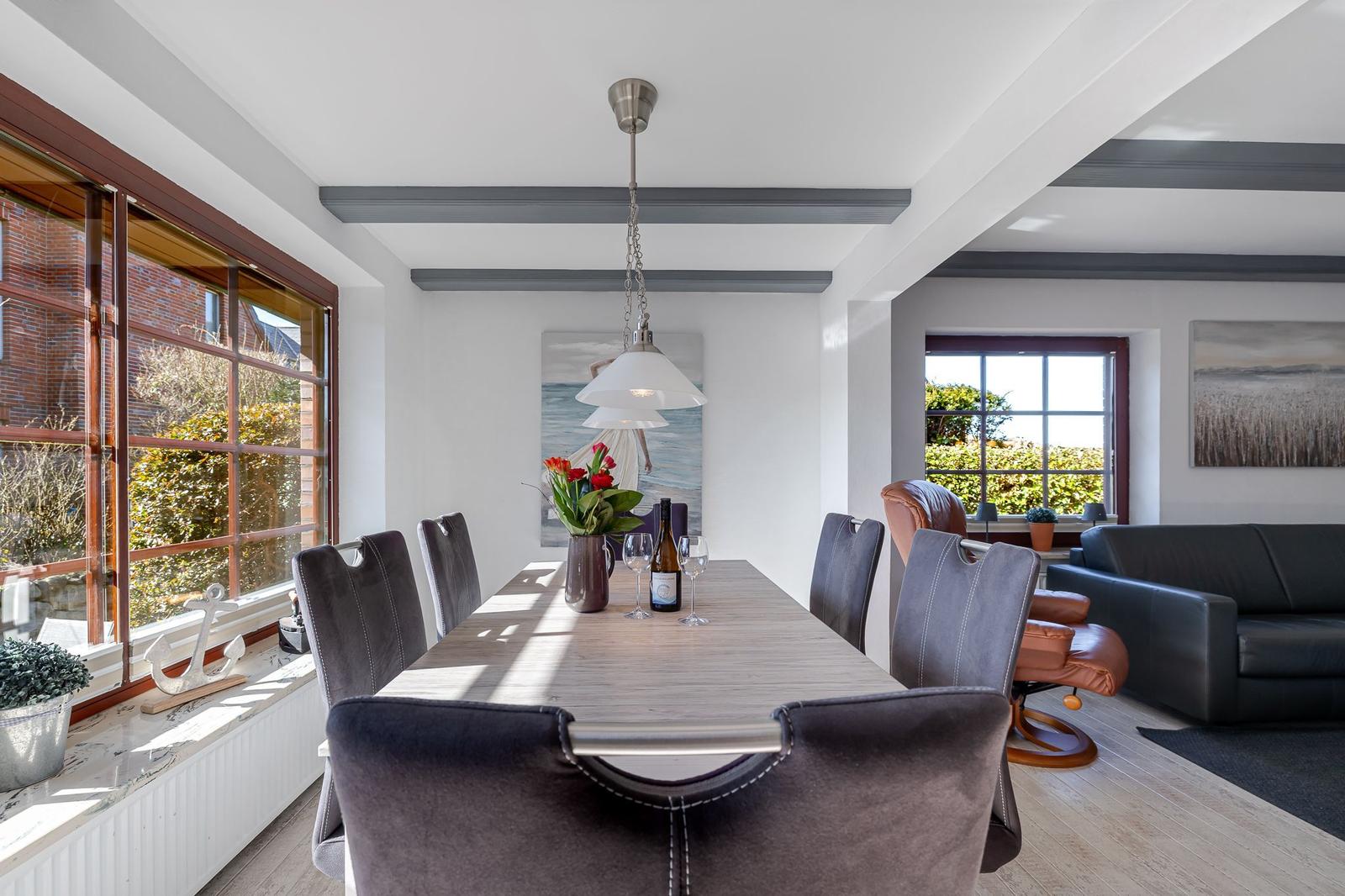 Esszimmer mit langem Tisch, grauen Stühlen und großen Fenstern. Wohnbereich mit Sofa und Sessel.