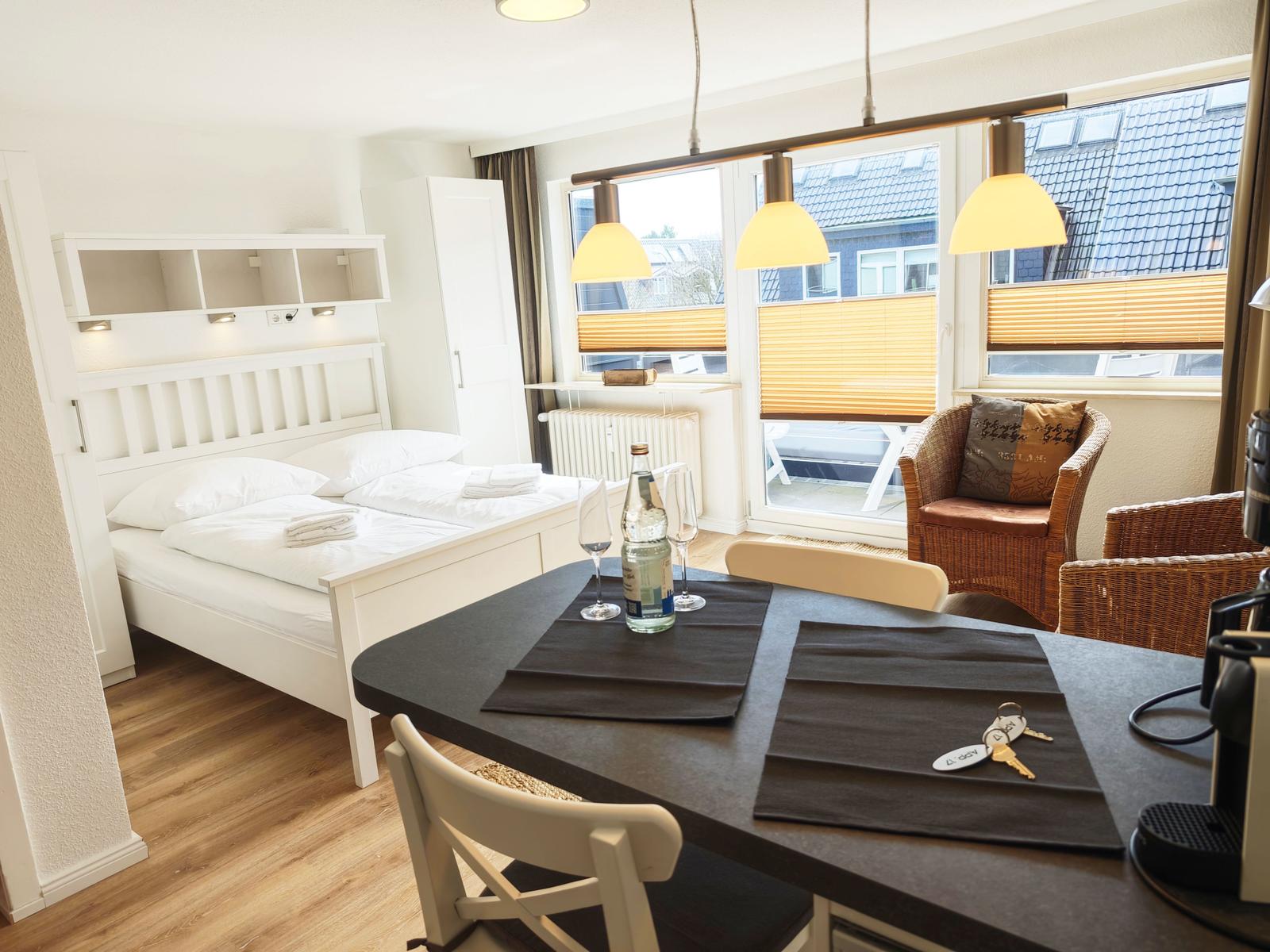 Schlafzimmer mit Bett, Tisch und Sessel. Fenster mit Jalousien und Lampe.