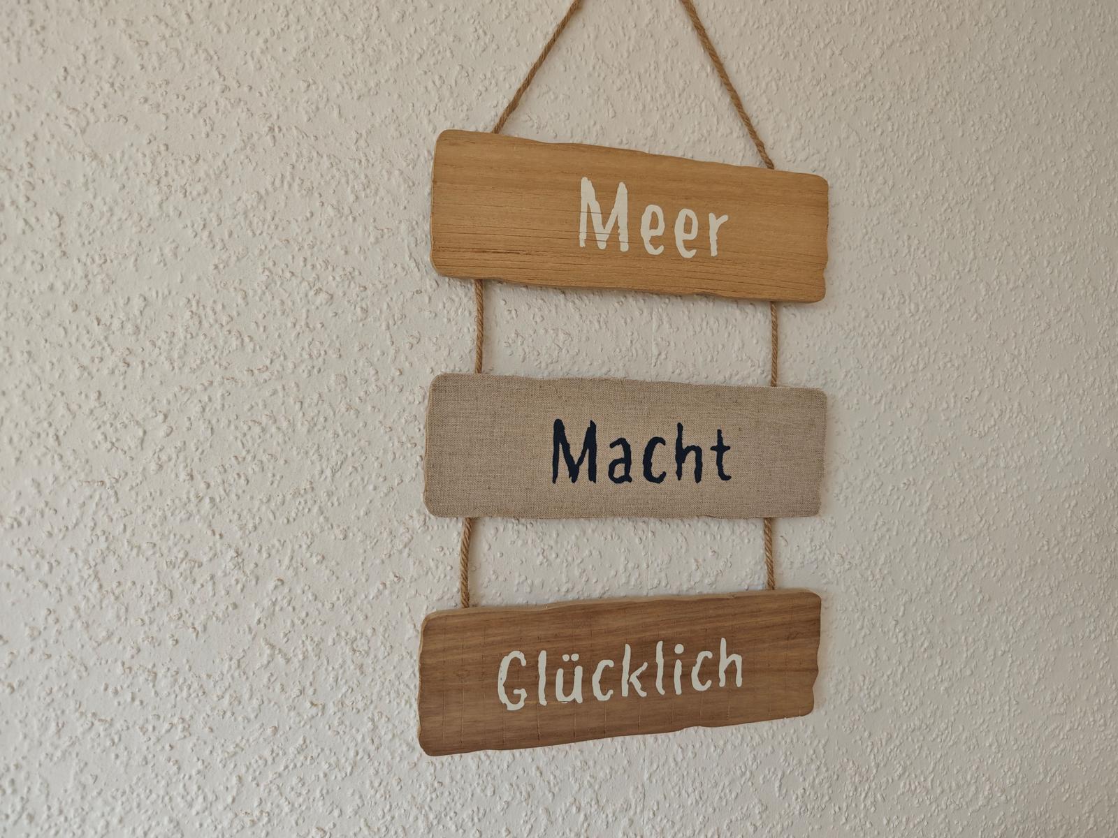 Drei Holztafeln hängen an einem Seil an der Wand. Sie zeigen die Worte Meer, Macht und Glücklich.