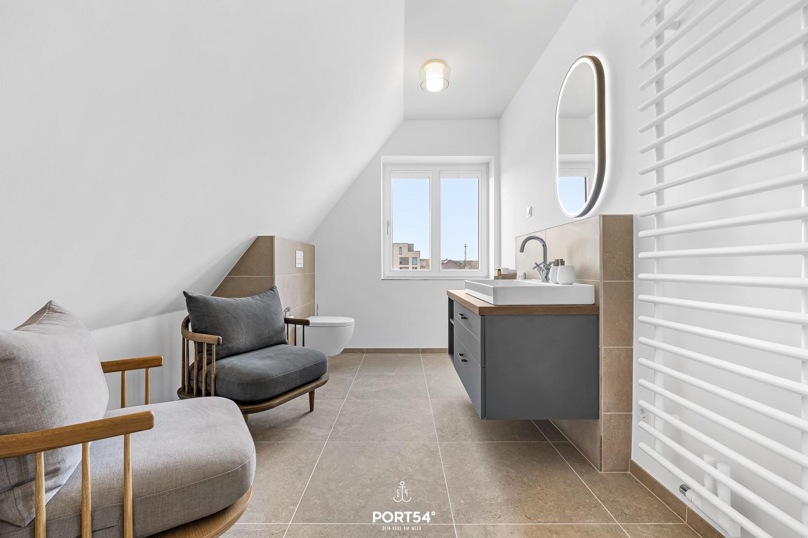 Badezimmer mit Waschbecken, Toilette, Fenster und Sitzgelegenheit.