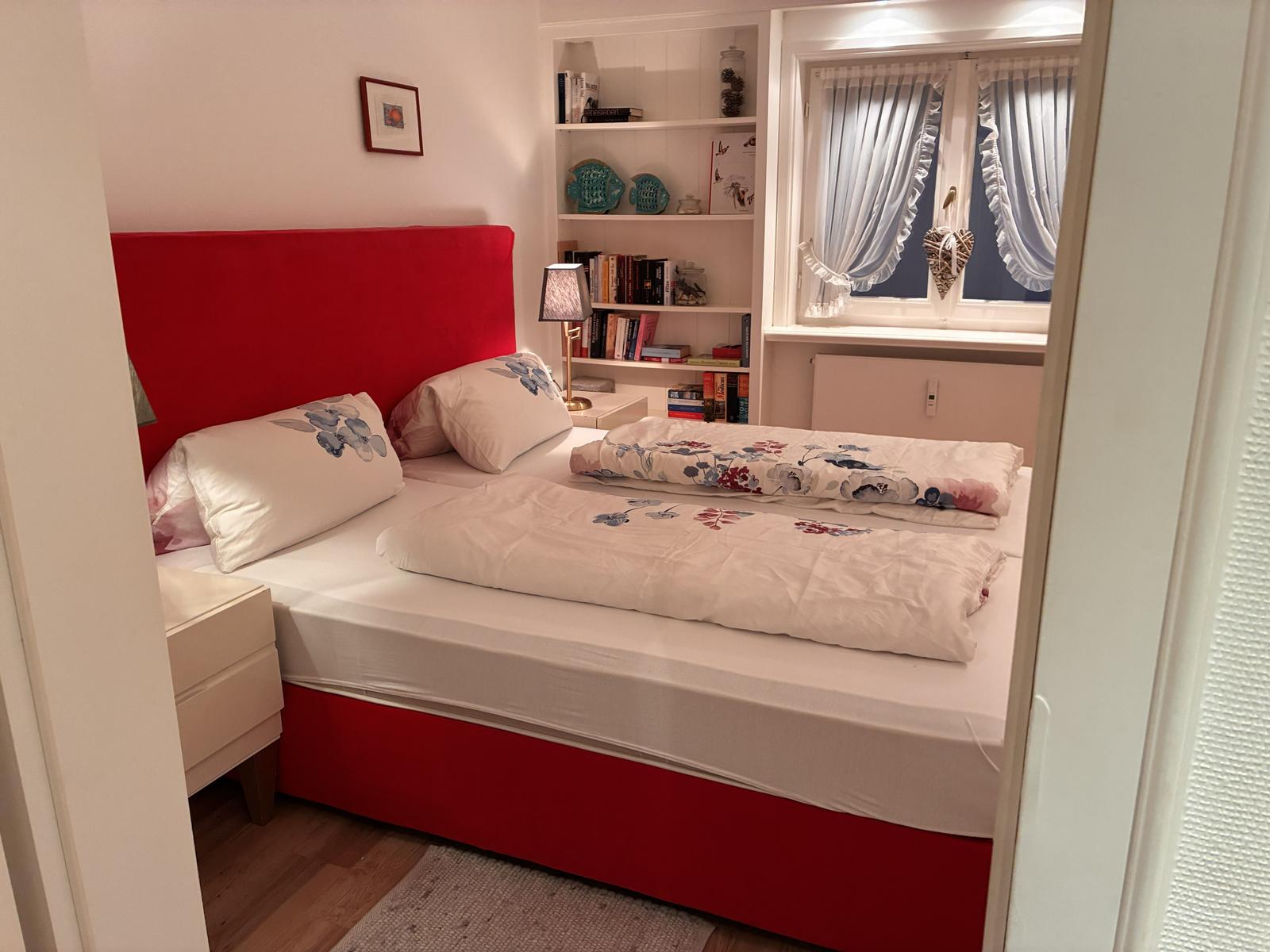 Schlafzimmer mit rotem Bett, Bücherregal und Fenster mit Vorhängen.