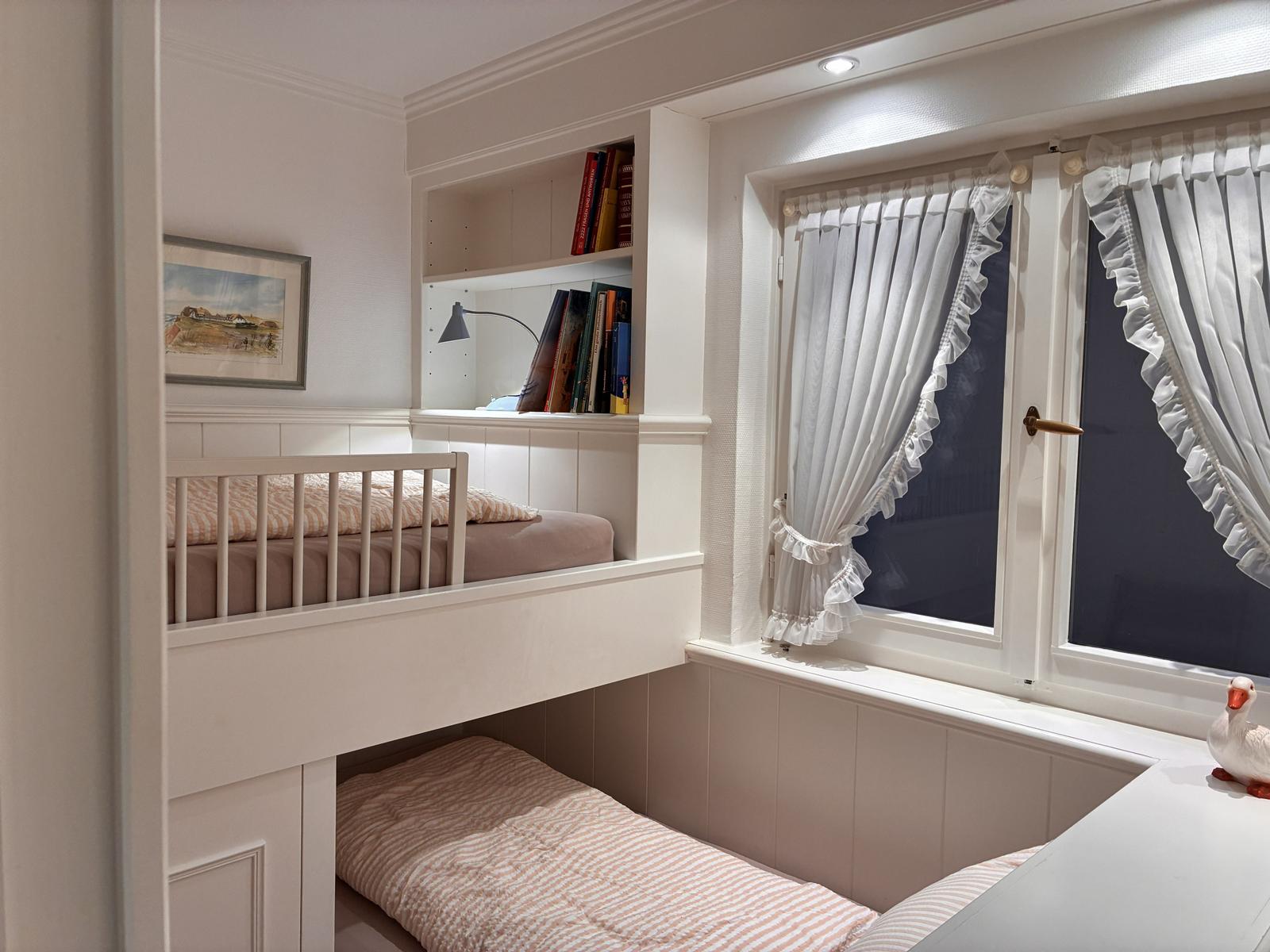 Zimmer mit Doppelstockbett, Bücherregal und Fenster mit weißen Vorhängen.