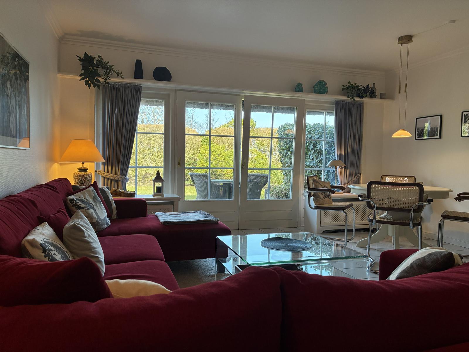 Ein Wohnzimmer mit rotem Sofa, Glas-Tisch und Fenstern zum Garten.