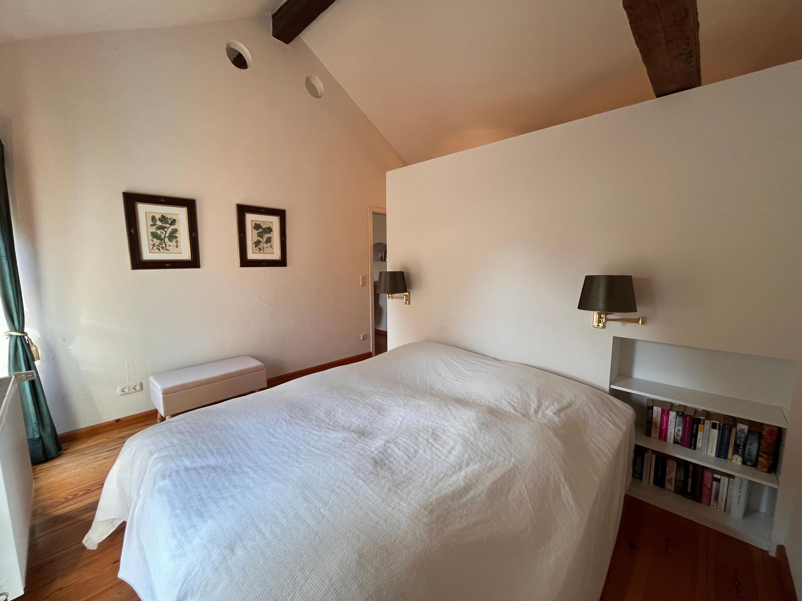Schlafzimmer mit Bett, Bücherregal und Wandlampen. Holzboden und schräge Decke mit Balken.