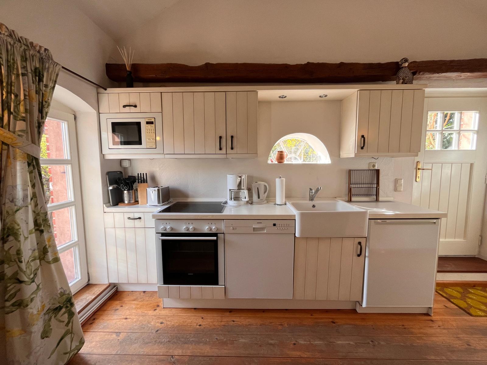 Küche mit weißen Schränken, Steinplatten und Holzboden. Fenster mit Blumenvorhängen.